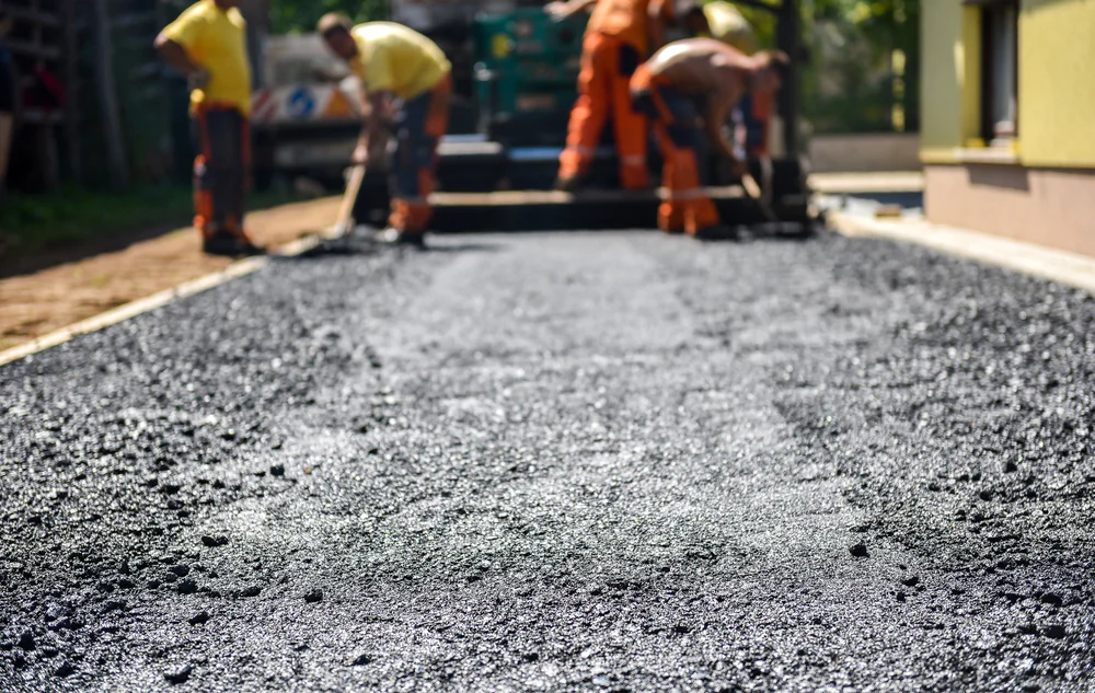 Workers in bright safety clothing lay fresh asphalt on a road, following recent commercial excavation Virginia. The foreground shows the textured, newly laid surface, while the team works with smoothing tools slightly out of focus in the background.