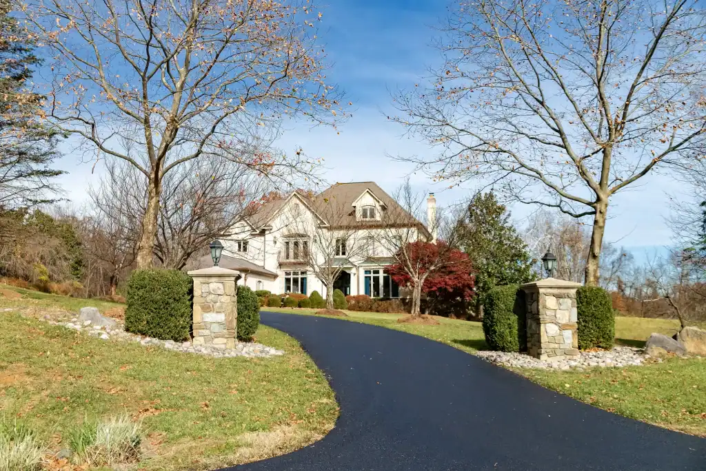 A large, two-story house with beige siding and stone accents sits at the end of a curved, paved driveway—evidence of expert residential excavation Virginia—surrounded by manicured lawns, shrubs, trees, and stone lamp posts under a clear blue sky.