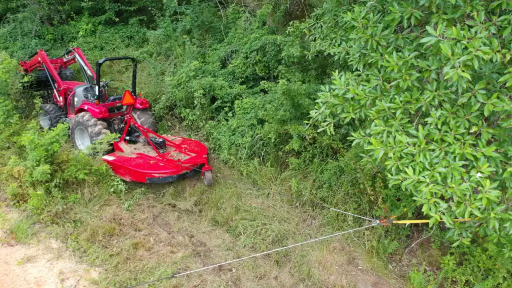 A red tractor with a mower attachment is stuck in tall grass near dense green bushes, secured by a cable tied to a nearby tree, as part of a land clearing Virginia project attempting to free it.