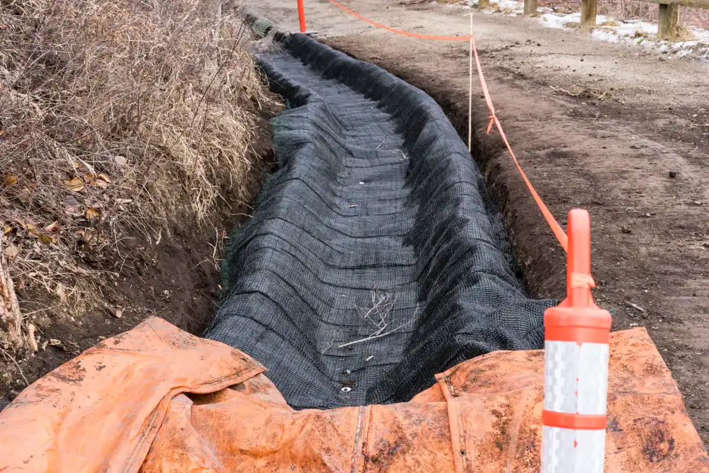 A shallow ditch along a dirt road lined with black erosion control fabric, bordered by orange safety tape and a reflective post, with dry vegetation on one side—typical of sites prepared for residential excavation Virginia.