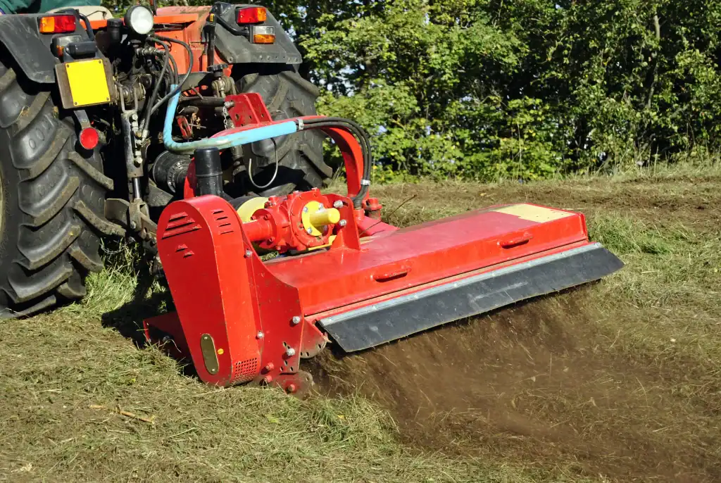 A red tractor-mounted mulcher processes grass and soil on a field, creating a dust cloud—an essential step in land clearing Virginia projects—with green trees visible in the background.
