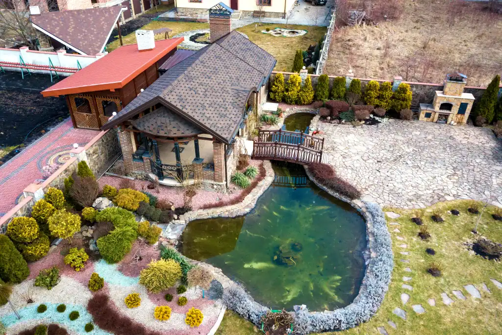 Aerial view of a landscaped backyard featuring a pond with a small bridge, stone pathways, various shrubs, trees, and a patio area next to a house with a brown roof.