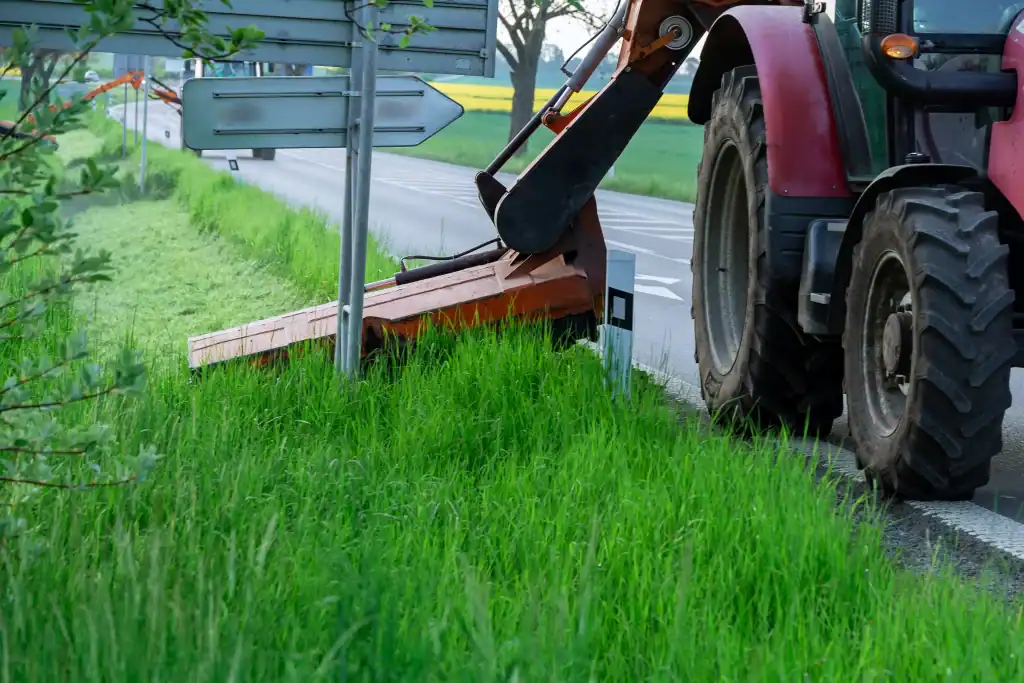 A tractor with a mower attachment trims tall grass along the roadside near traffic signs, with green fields and a partially visible paved road in the background.