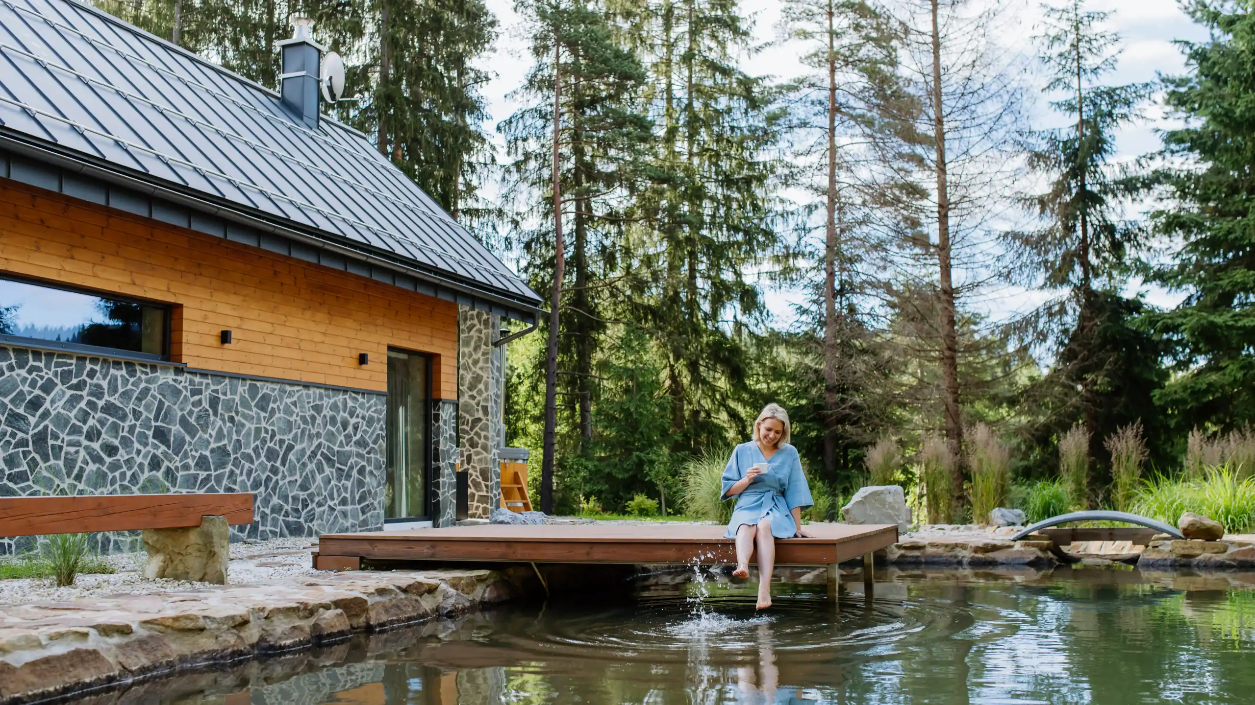 A woman in a blue robe sits on a wooden deck, dipping her feet in a pond beside a modern cabin surrounded by tall trees and lush greenery. She appears to be reading or writing.