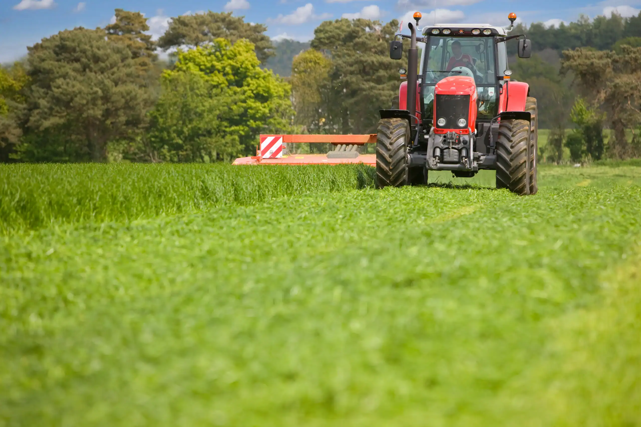 A red tractor drives across a lush green field under a blue sky with scattered clouds, surrounded by trees in the background.