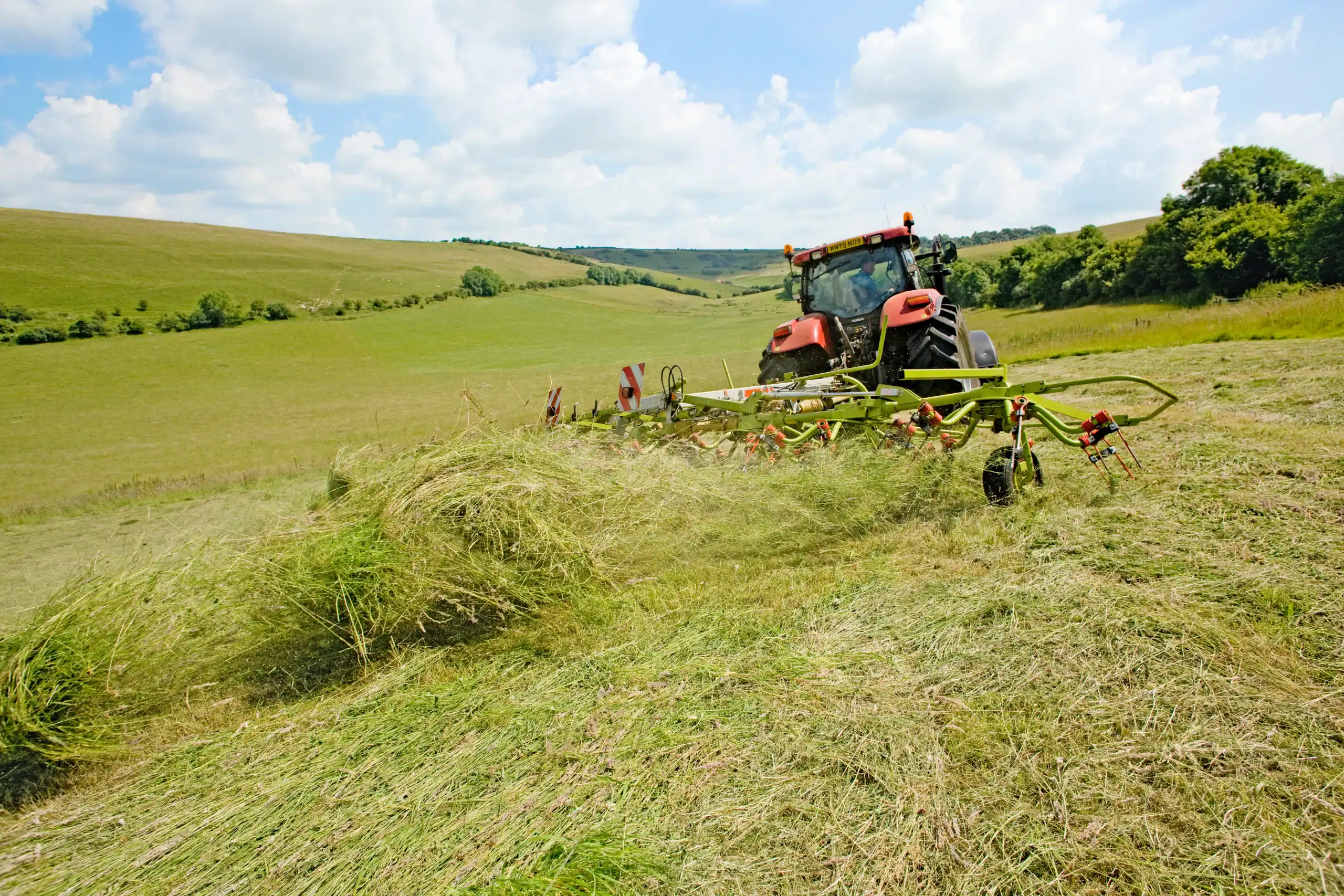 A red tractor with a hay rake attachment is working in a large, grassy field under a partly cloudy sky, gathering and turning hay with green hills and trees in the background.